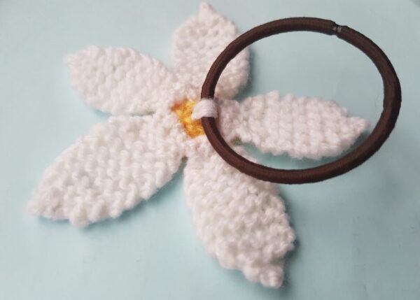 Close-up of a handmade white knitted daisy attached to a brown elastic hair bobble on a light blue background.