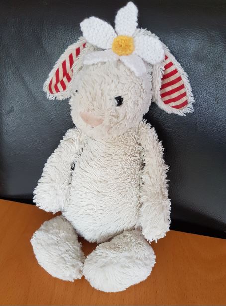 Cream-coloured soft toy bunny with red striped ears wearing a knitted white daisy flower on its head, seated against a black background.