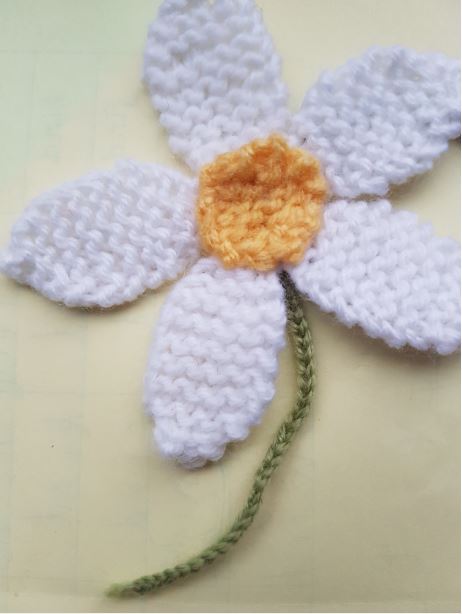 Completed hand-knitted white daisy with a yellow centre and green embroidered stem on a pale background.