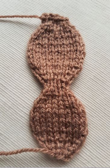 Knitted brown double-lobed shape resembling a figure-eight, laid flat on a textured cream fabric, likely used for a reindeer’s body or head component.