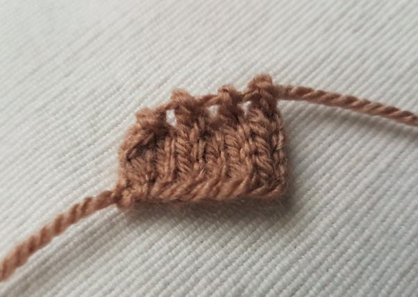 Small rectangular knitted brown piece with evenly spaced stitches, likely part of a reindeer ear or facial detail, displayed on a cream fabric background.