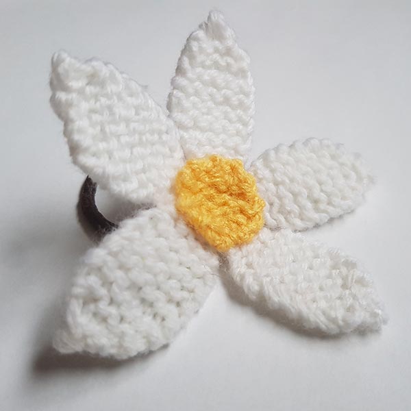 Handmade white knitted daisy with a yellow centre, attached to a hair elastic, displayed on a white background.