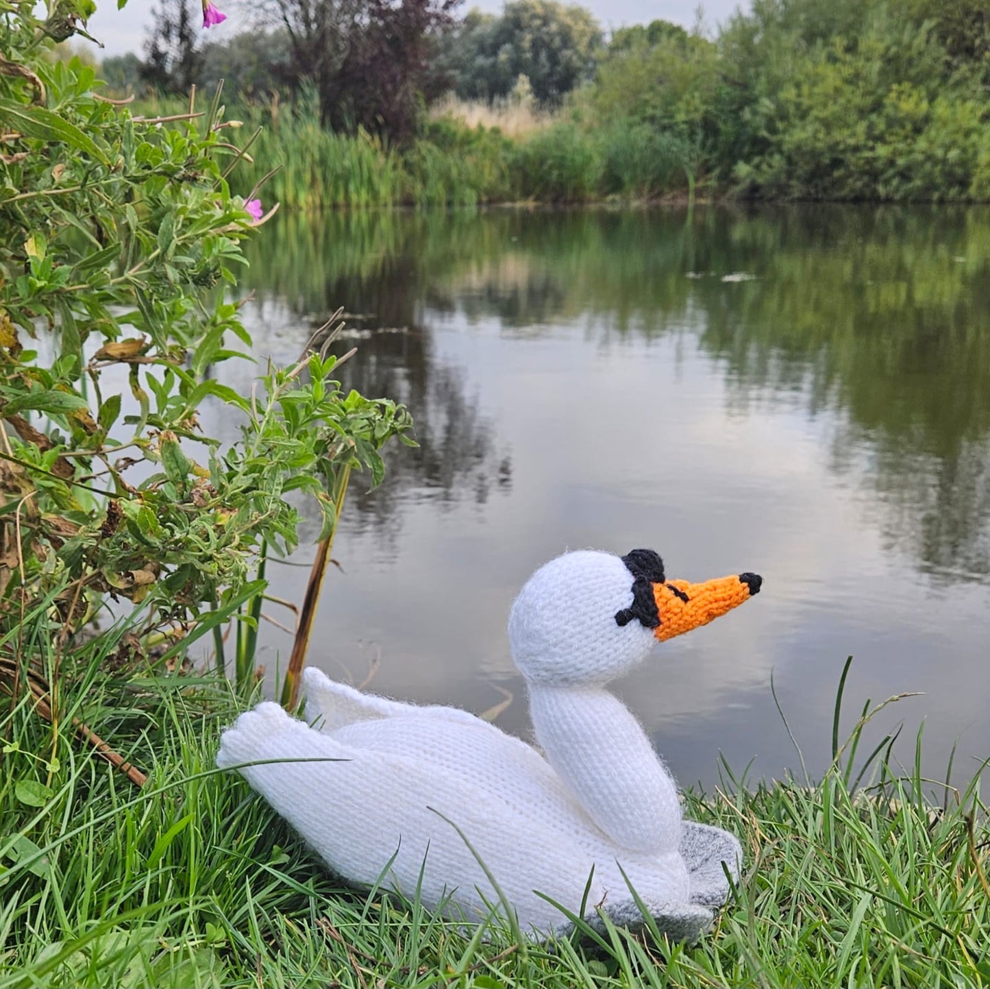 Knitted swan toy on a grassy bank by a pond with trees in the background