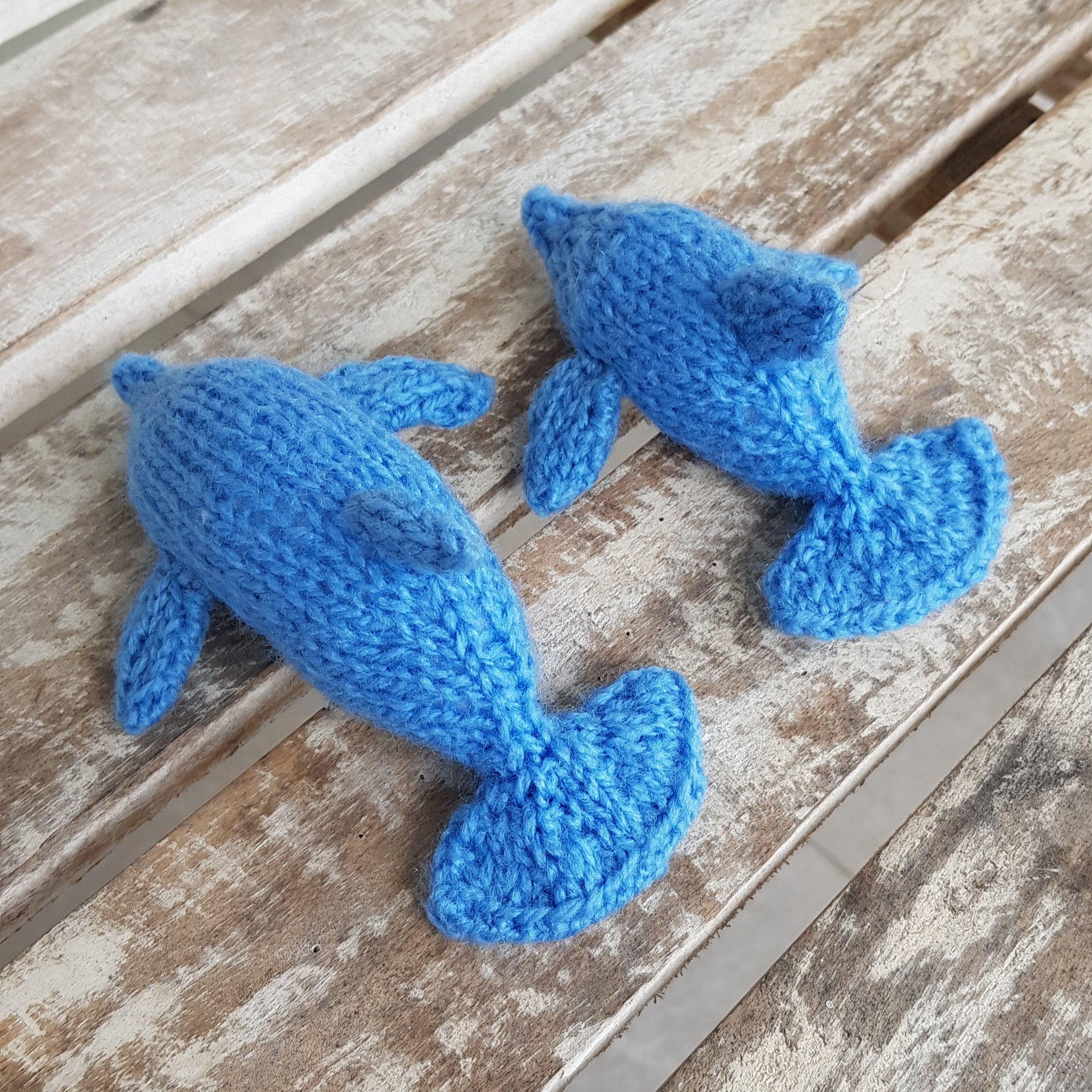 Side view of knitted dolphin toys – large and small – handmade from beginner-friendly dolphin knitting pattern, displayed on rustic wooden bench, by Fitting in Knitting.