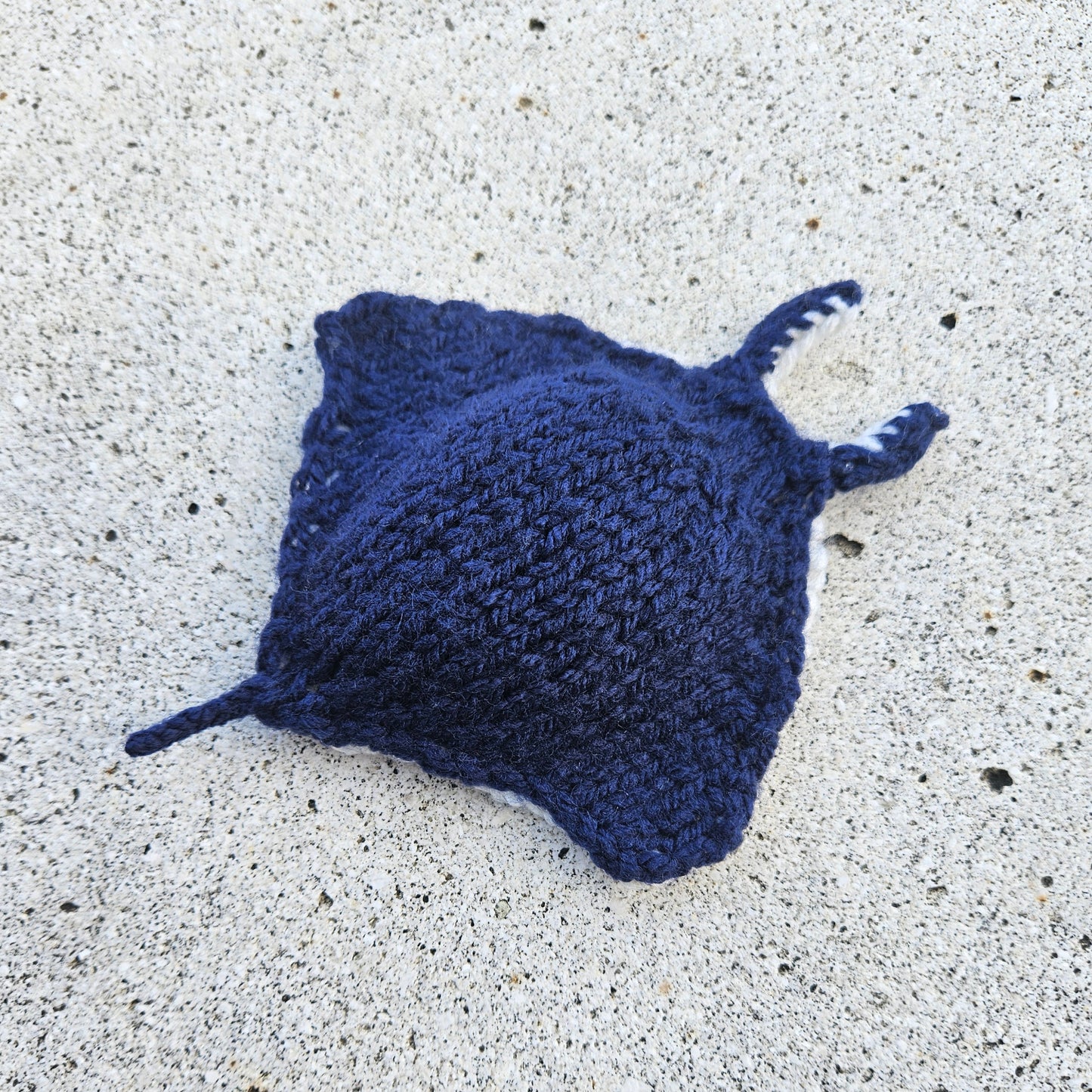 Dark blue hand-knitted manta ray toy basking on golden sand, with stitched details.