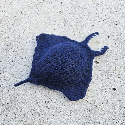 Dark blue hand-knitted manta ray toy basking on golden sand, with stitched details.