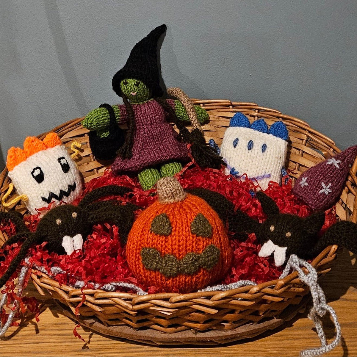 A collection of handmade, knitted Halloween toys arranged in a wicker basket filled with red shredded paper.