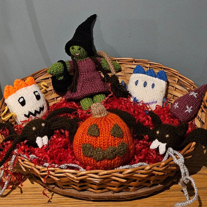 A collection of handmade, knitted Halloween toys arranged in a wicker basket filled with red shredded paper.