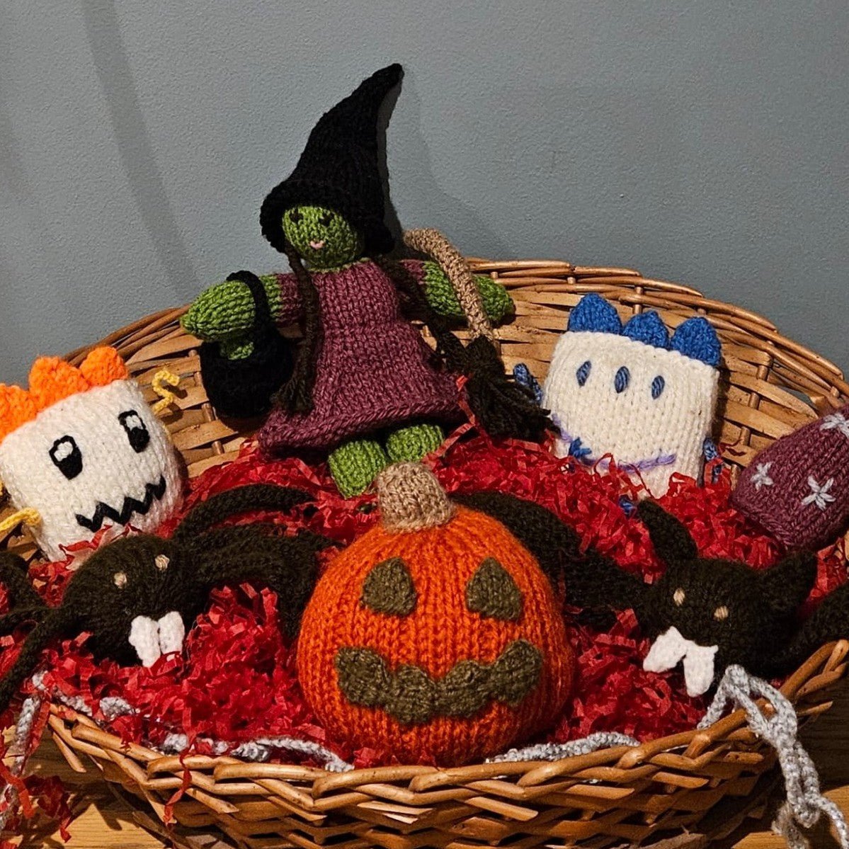 A collection of handmade knitted Halloween toys arranged in a wicker basket filled with red shredded paper.