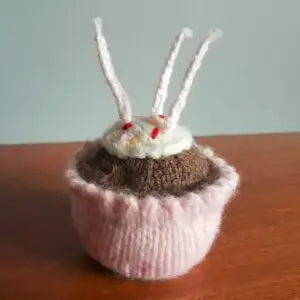 Knitted cupcake with pink frosting, brown base, and white candles on a wooden surface.