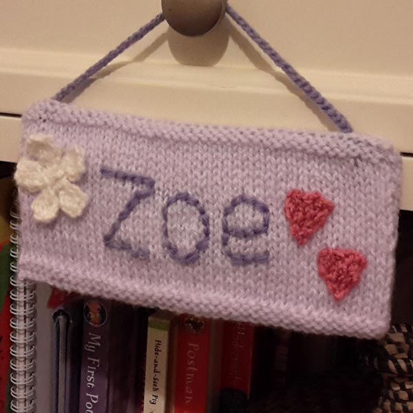 Hand-knitted lavender name sign with the word “Zoe” stitched in purple yarn, featuring a white flower and two pink hearts, hanging from a matching knitted loop.
