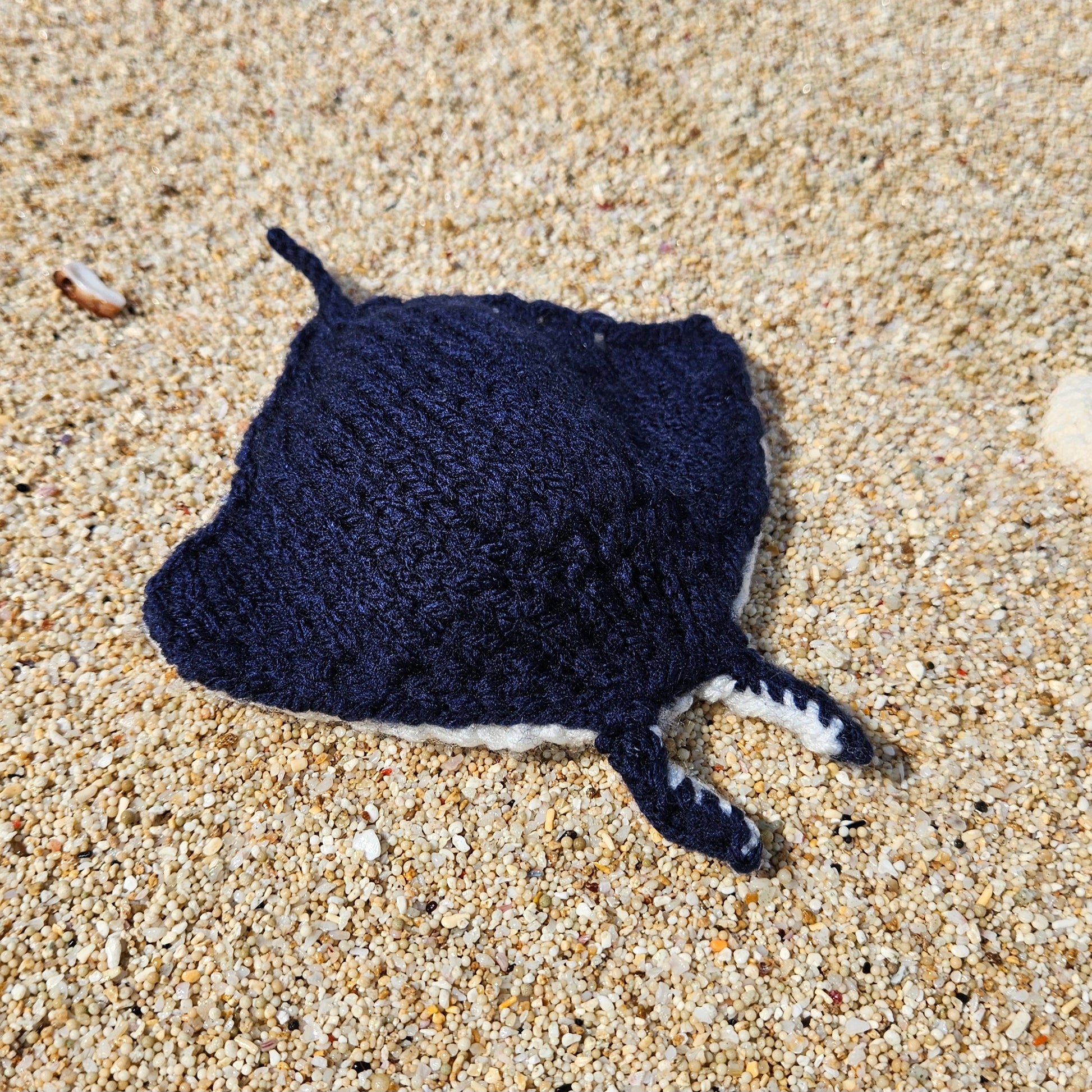 Dark blue hand-knitted manta ray toy resting on the sand, part of the Fitting in Knitting collection.