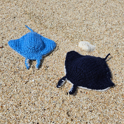 Two knitted manta ray toys, one dark blue and one light blue, side by side on the beach.