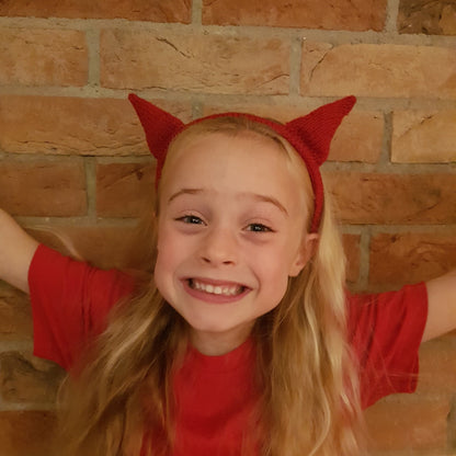 A young girl with long blonde hair smiling at the camera, wearing a red devil horn headband and a red T-shirt.