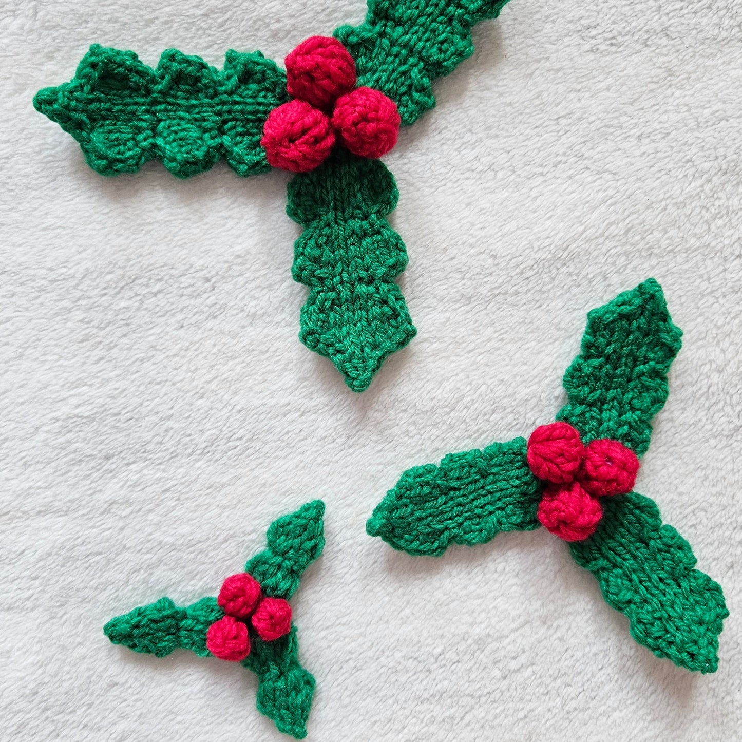 Three green knitted holly leaf ornaments with red berries, arranged on a white textured fabric background.