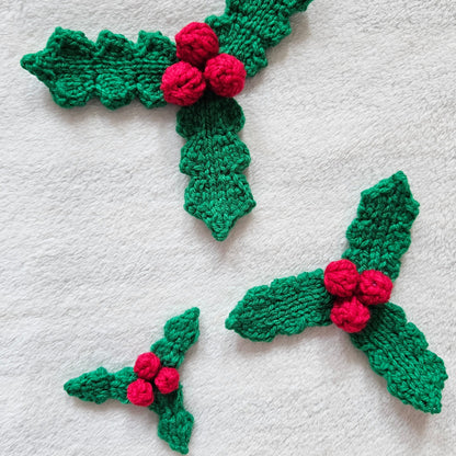 Three green knitted holly leaf ornaments with red berries, arranged on a white textured fabric background.