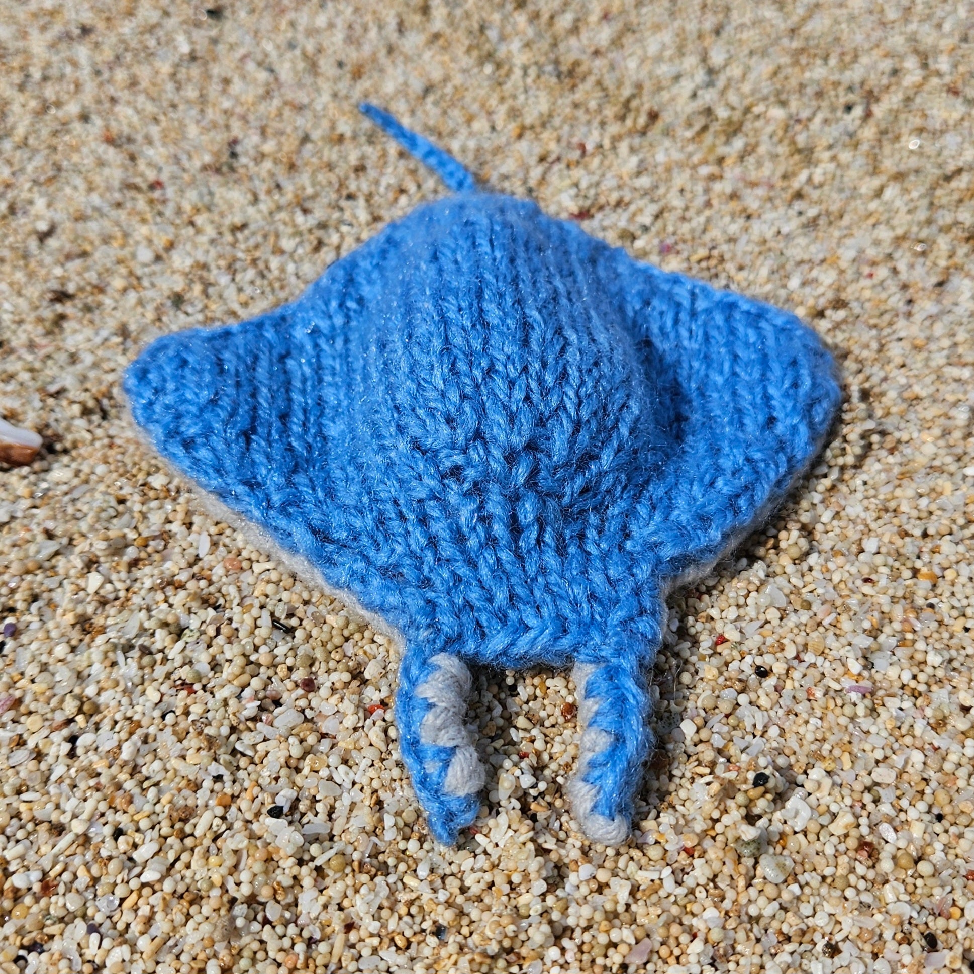 Light blue knitted manta ray toy placed on a sunlit sandy beach.