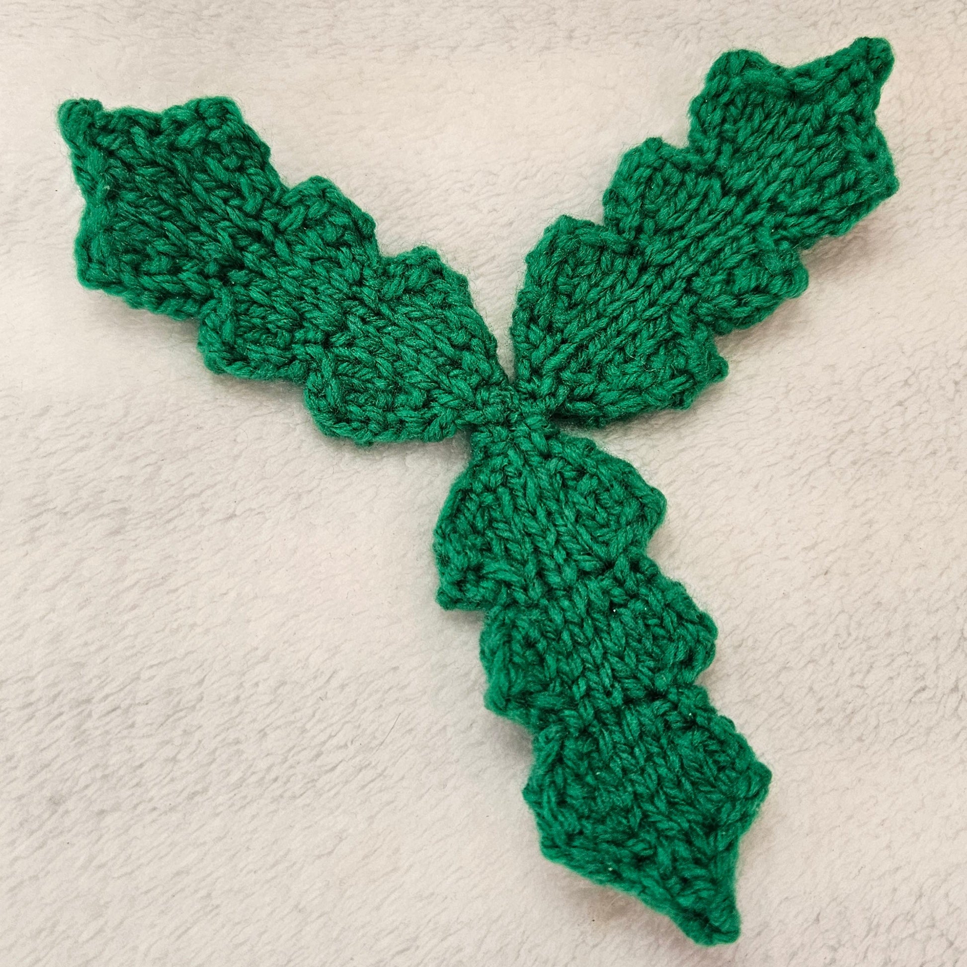 A green knitted three-petal holly arrangement, used a decorative applique or craft embellishment, laid flat on a textured off-white fabric background.