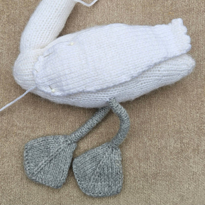 Partially assembled white knitted swan with a narrow wing pinned to its body and grey legs and feet.