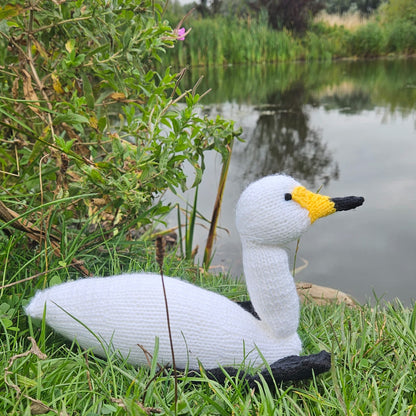 Knitted swan toy on grass near a pond with greenery