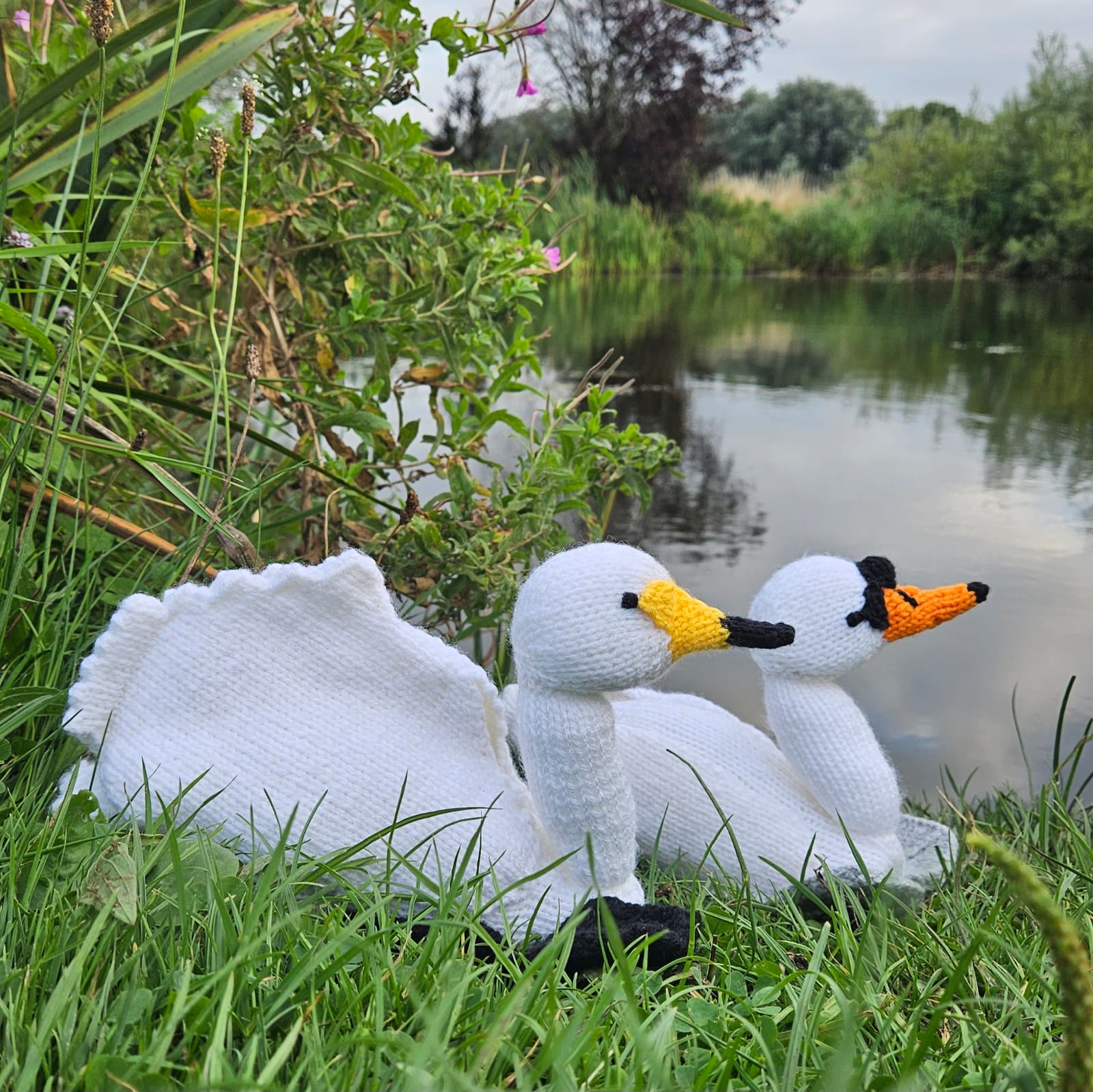 Knitted swan toys on grass near a pond with greenery