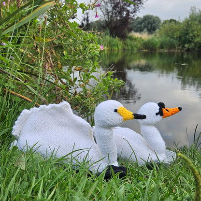Knitted swan toys on grass near a pond with greenery